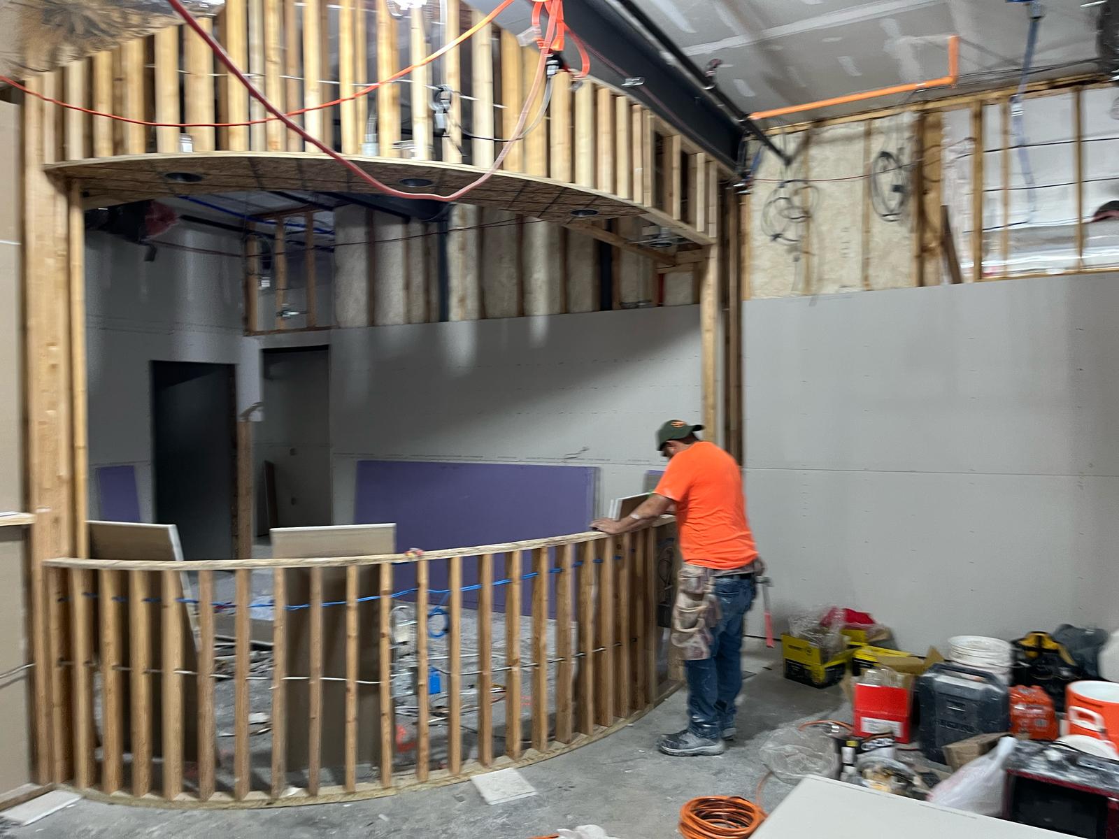 Construction worker in orange shirt working on framing installation in a partially finished interior space, featuring drywall, tools, and materials for framing and drywall services.