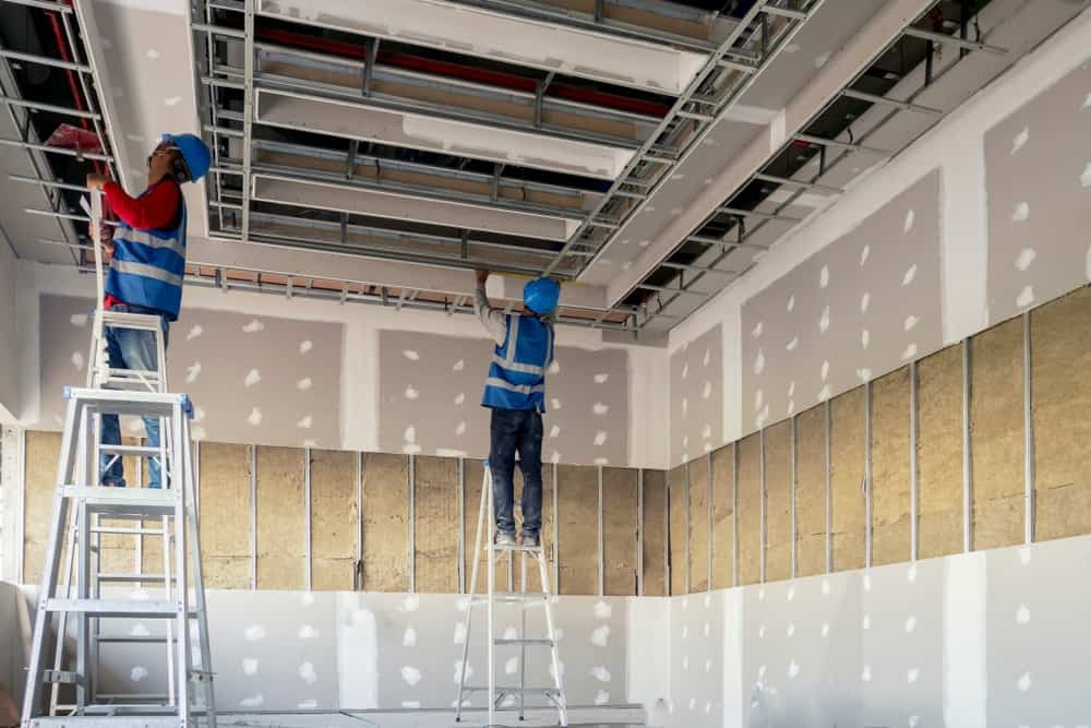 Two construction workers on ladders installing drywall and framing in a partially finished interior, showcasing craftsmanship in drywall installation.