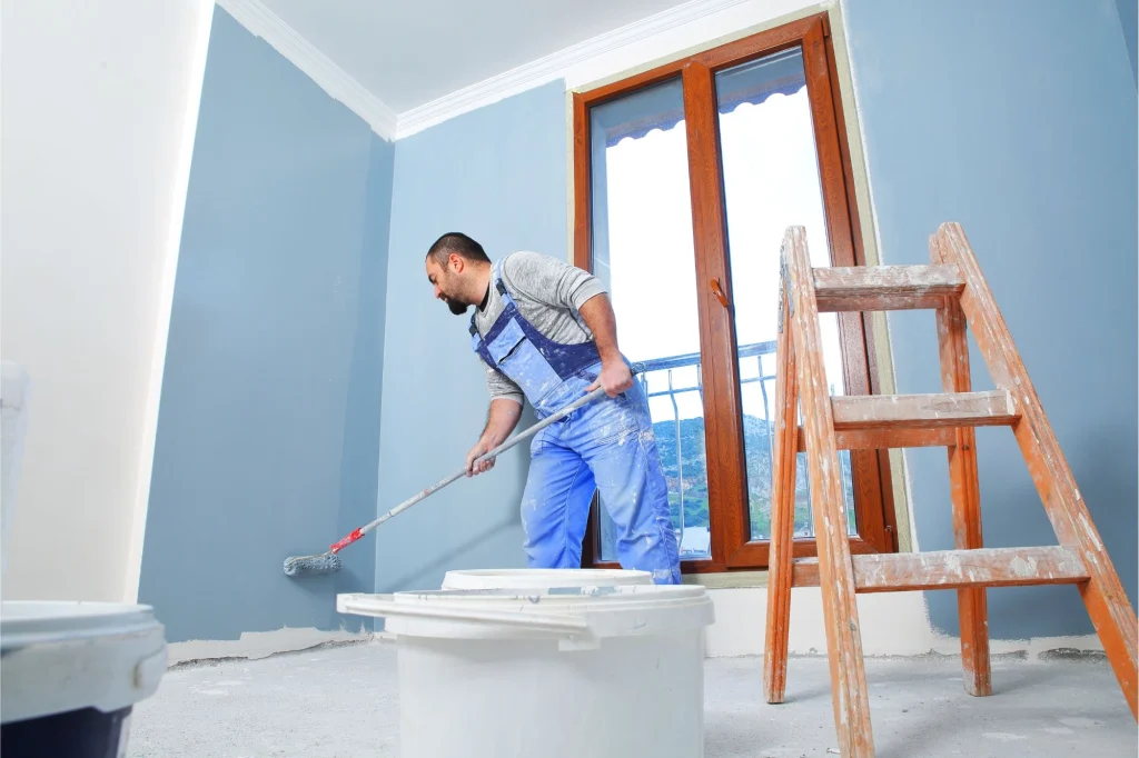 Man painting a blue wall with a roller in a residential interior, featuring a wooden ladder and paint buckets, showcasing professional interior painting services by Framing to Drywall.